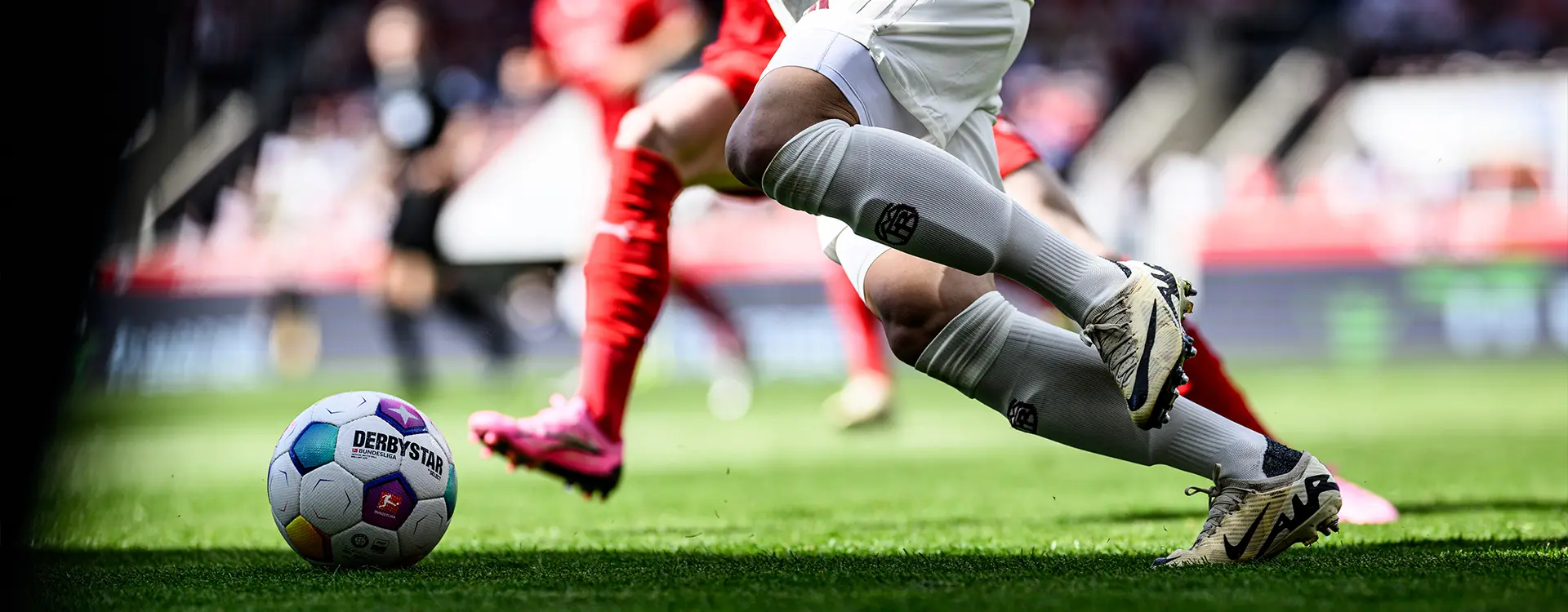 The image shows a soccer player, who appears to be looking down at the ball with attention. They are wearing a jersey that matches their team's colors and have a foot forward as part of their run or kick. The player is mid-motion, and the focus of the photograph is on them from this angle. In the background, there is another player and what might be a goal goalpost to the right. The setting suggests an active soccer match with two teams engaged.