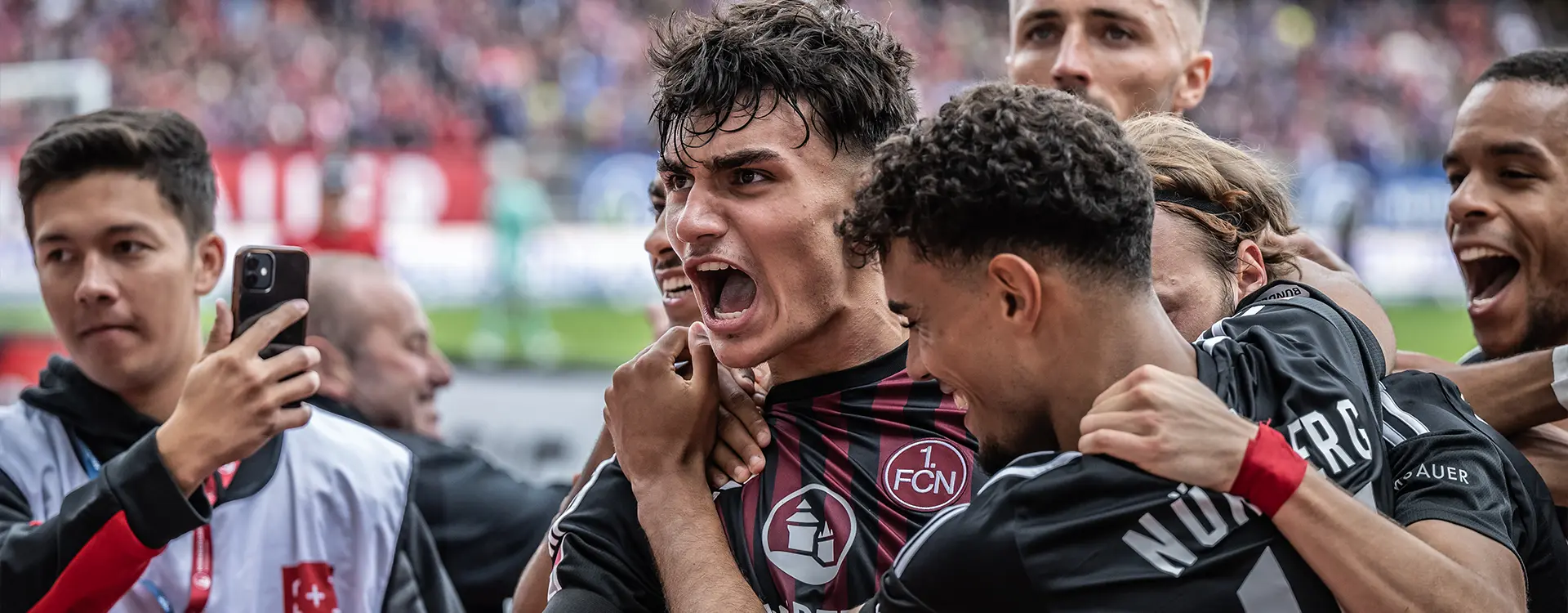 Das Bild zeigt eine Fußballmannschaft in einem Stadion. Die Spieler stehen zusammen auf dem Rasen, feiernd einen Sieg oder ein Erfolg und zelebren während einer Pause. Einige der Spieler halten vor sich ihr Trikot und feiern durch gesteckte Fingern und eine erhobene Stimmung. Manche der Spieler nutzen die Zeit, um ihre Zangen zu spitzen oder den Trinkgeber mit ihrem Trikot anzugiechen. Hinter den Spielern gibt es Fans, die entgegen die Regel auf den Rasen gekommen sind, sich jedoch nicht direkt beim Team einmischen können. Zwei Fans haben ihre Smartphones gezogen und sind so beschäftigt, dass sie das Ereignis der Spieler nicht ausfällig beobachten können.