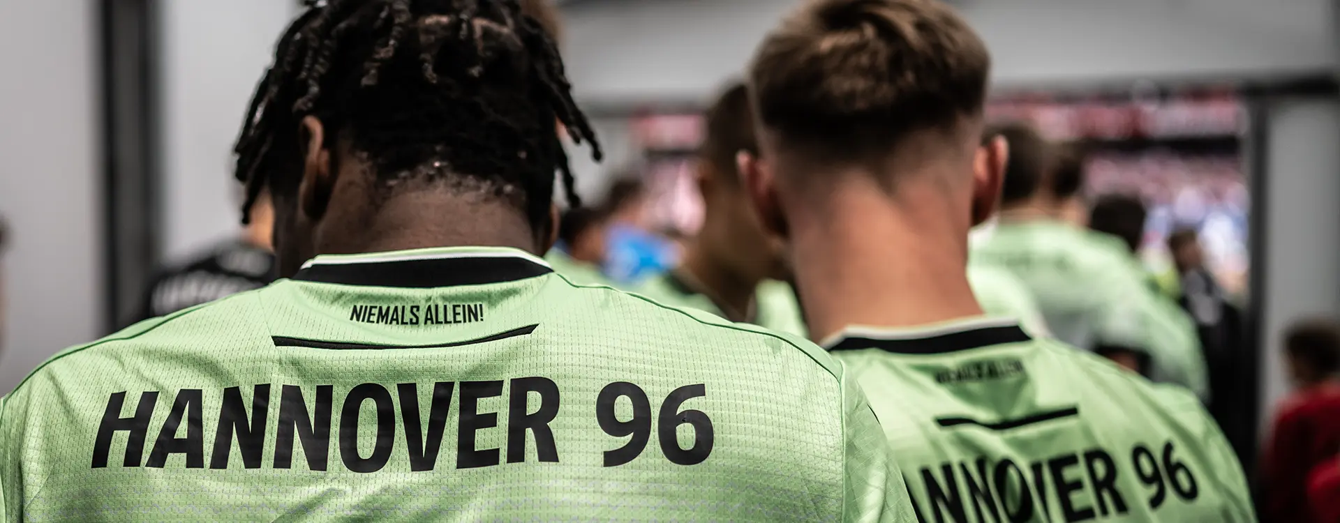 The image depicts a group of individuals, likely athletes, wearing matching sports jerseys with the name "HANNOVER" and the number "96." These jerseys suggest that this is a football match related to Eintracht Hannover, which could imply that they are members of the team's management or staff. The photo appears to have been taken indoors after an event, as indicated by the environment in the background and the expressions on their faces, possibly reflecting exhaustion from the game or a meeting that has just concluded.