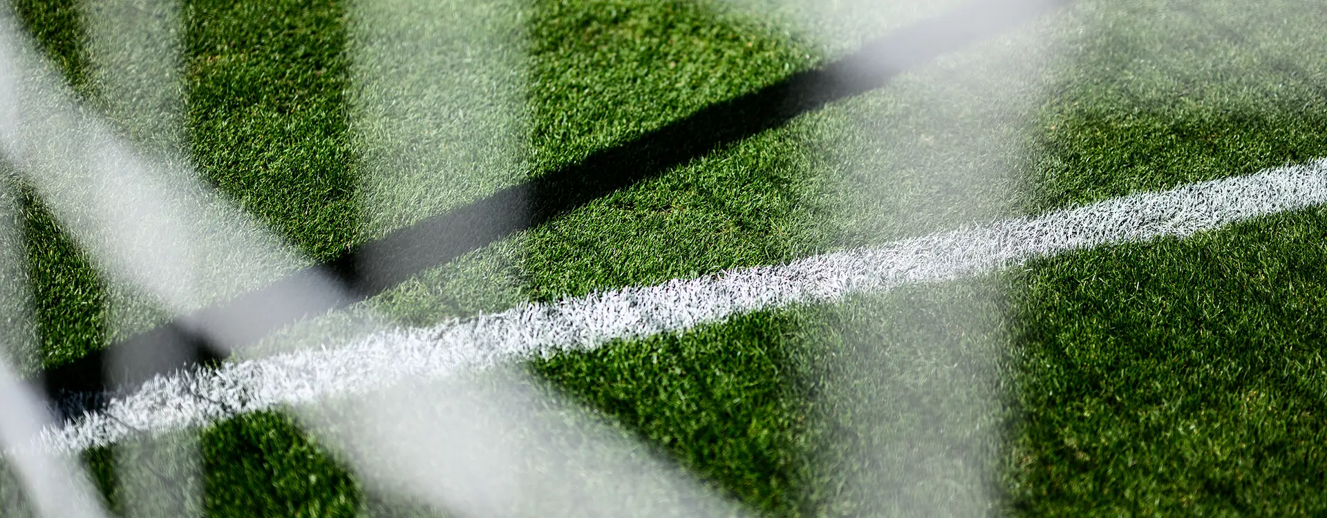 The image shows a close-up of a soccer goal net with netting, likely due to its protective nature. In the background, there's a blurred view of what seems to be a field marked with white lines, possibly indicating sporting activity or an area designed for sports. There are also two football posts visible in the lower right corner of the image, standing outside of the soccer goal. The photo appears to have been taken from behind the netting, and it's taken vertically, giving its perspective.