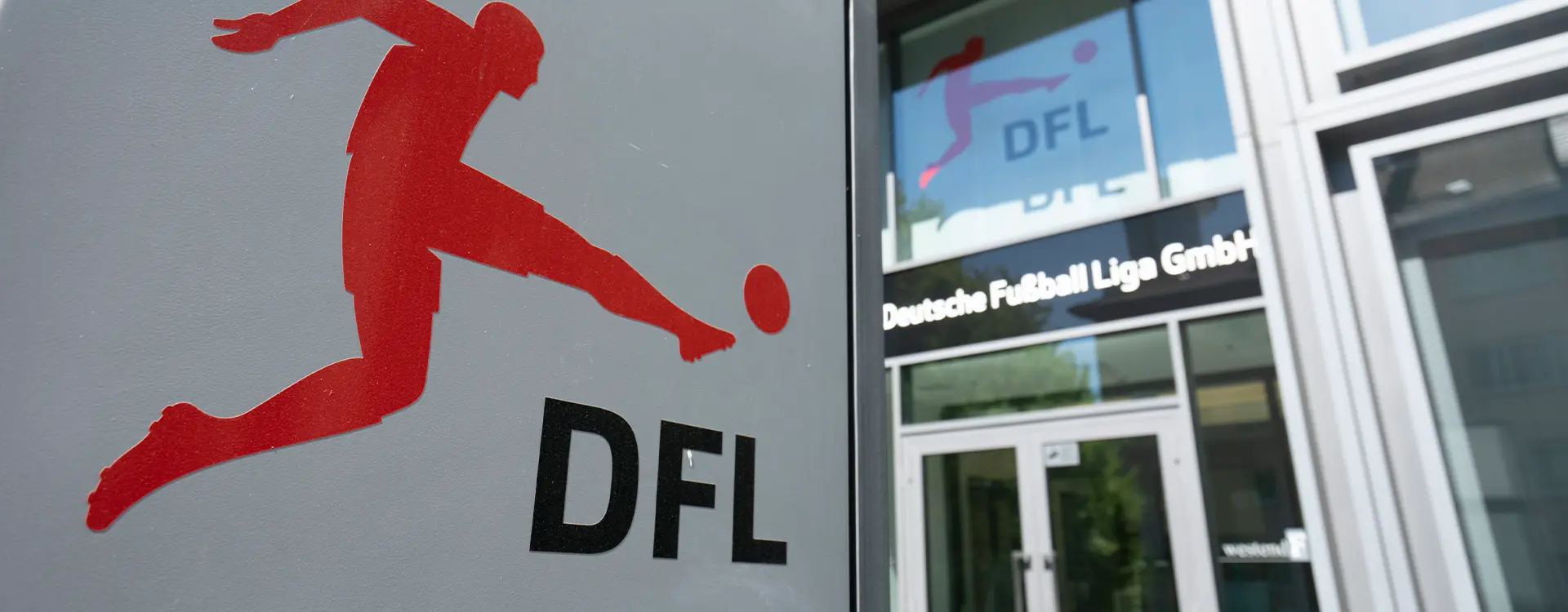 In the image, you can see a sign with a red silhouette of a soccer player that appears to be kicking a soccer ball next to the word "DEFI," which is likely short for "Definitivo." Additionally, there's text in Polish underneath the logo, but I'm unable to provide translations. Next to the sign is an entrance to a building, and on a glass panel within the entrance, you can see the names "Marcin Kwaśniewski" and "Olga Michanowska." The background suggests that this image was taken during daylight, as there is natural light.