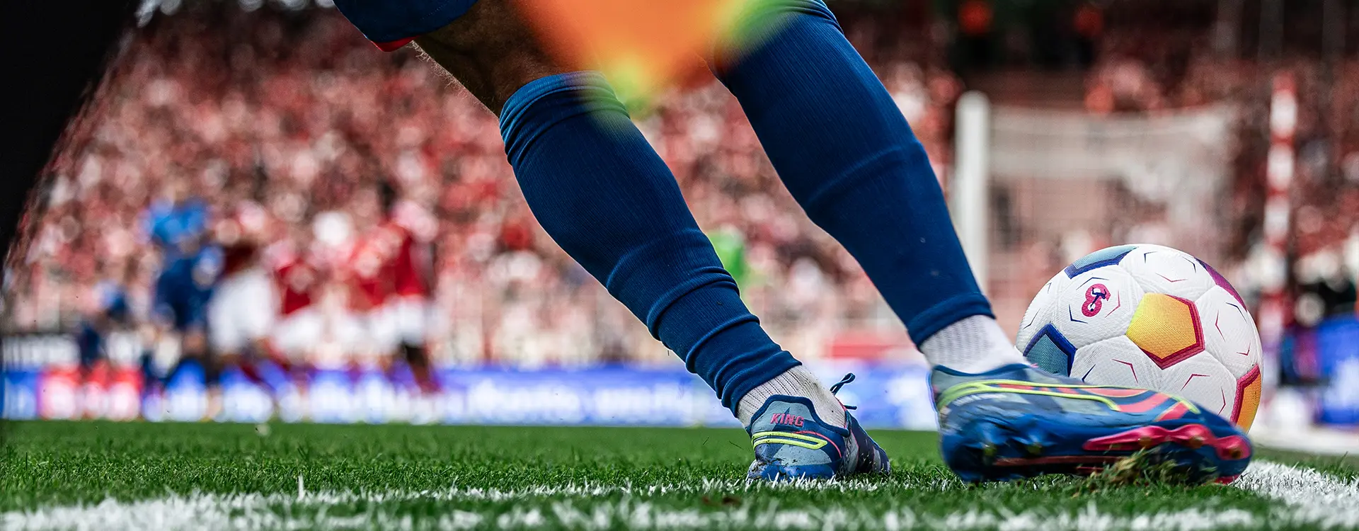 The image you provided is a promotional or advertising image for a football (soccer) match, showcasing a player in action kicking a ball on a pitch. The focus is on the sport and the atmosphere of the game. In the background, there are spectators visible through the goalposts. There is no text present on the image to provide additional context or information about the event or players involved.