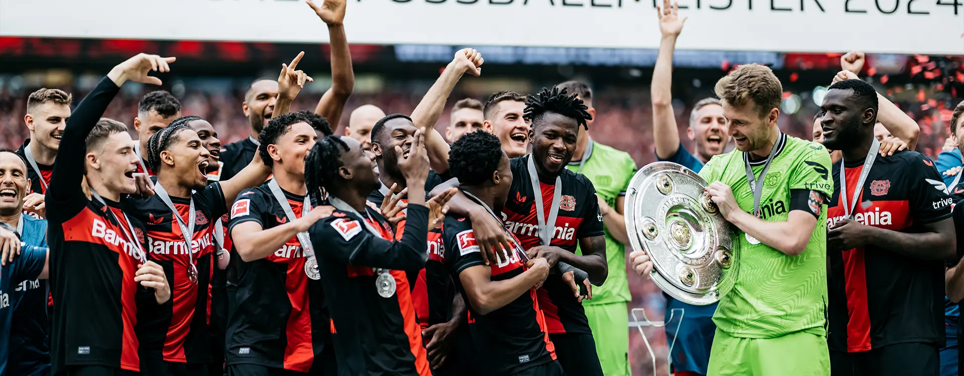 In the image, there is a moment being captured at what appears to be an awards ceremony or end-of-season festivities for a football (soccer) team. A group of players, distinguished by their jerseys and medals around their necks, are congregated together in celebration. Some players are giving the impression of applauding, while others have their arms raised in a gesture that could be interpreted as victory or jubilation. There is one player who stands out from the others; he is standing slightly apart from the group and appears to be holding up his trophy with a mix between relief and joy. The team's name on his jersey, "Chelsea," suggests it might be the Chelsea football club.
The photo captures this moment with the people in focus, and there are banners in the background that feature what seems to be sponsor or event logos, contributing to the festive atmosphere of the occasion. The text on the left side of the image is very small and not clear enough to be read.