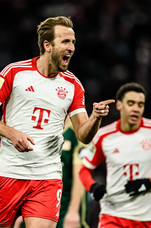 The image shows a soccer player on the field during what appears to be a game. He is wearing a FC Bayern Munich jersey, which is predominantly red and white with blue accents and number nine in gold. The player has his mouth open and seems focused on something or someone off-camera as his gaze does not match any specific object in the frame. His posture suggests he has just completed an action like kicking the ball or celebrating and he's turned to look back at someone or something, possibly towards opponents or teammates. The player's expression is intense, reflecting concentration and determination. In terms of the environment, there are stadium lights in the background, indicating that the game is taking place during evening or night hours. The image itself has been digitally altered or overlaid with a watermark or logo, as evidenced by the presence of text and symbols on the photo, which would typically indicate copyright or ownership. Please note that this description is based on the visible content in the image and does not include any speculative or non-specific elements.