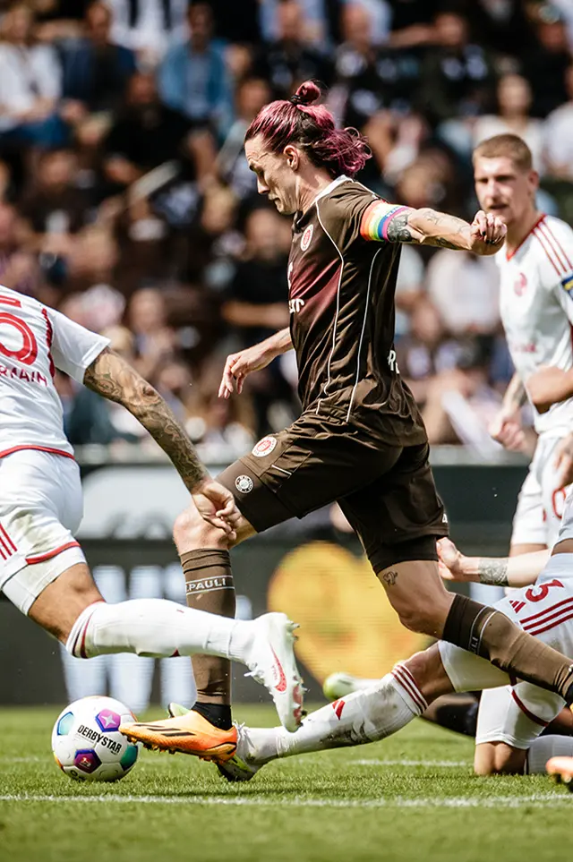 The image is a collage and consists of two separate photographs related to soccer. In the top half, we see a scene from a professional soccer match. A player wearing a brown jersey with white stripes is running towards the camera with an intent look on his face, holding possession of the ball, while another player in a red jersey with white trim and black shorts is in pursuit. The action photo captures the dynamic movement typical of a soccer game. In the lower half, on the right side of the image, there is a color photograph of a female soccer player with short brown hair wearing a purple top that matches her hair color. She appears to be playing for a team as indicated by the jersey with sponsor logos. Additionally, she has visible tattoos on her arms and is engaging in aggressive play, as evidenced by her kicking action toward another player's shins during the match. The context of the image suggests it may be intended as commentary on gender in sports or a comparison between amateur and professional athletes.