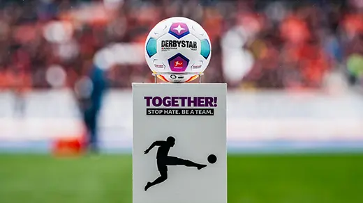 The image shows a sports ball mounted on a sign that says "BONJEAN PARK." The sign is standing at the sidelines of a rugby field, where a match has just concluded, as indicated by a large scoreboard behind it. In front of this ball on its stand, there's promotional material in multiple languages for the UEFA Euro Championship 2016, featuring the official mascot "Supervene" of that tournament along with a sign promoting France as the host nation. The image has a somewhat disjointed appearance, where different elements are pasted together into a collage, and it's not centered on any particular subject but rather a mix of these items.