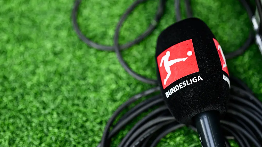 The image shows a microphone with audio cables connected to it, positioned on what appears to be a microphone stand, next to a printed cardboard "press" badge that is part of the stand. To left and right of the microphones are two black professional video cameras. In front of these items is a black microphone stand.
In the background, there's green astroturf with a watermark logo representing DFL (Deutsche Fußball Liga), which includes a soccer ball. Below the logo, it reads "Bundesliga", indicating this setup may be for a DFL press conference or similar event. On the right side of the microphone, there's an open black umbrella, commonly used for shielding media equipment from weather conditions.
The lighting on the microphones suggests they are connected to battery packs or chargers, which are placed in front of them on the floor. The overall setup is designed for a professional broadcast environment.