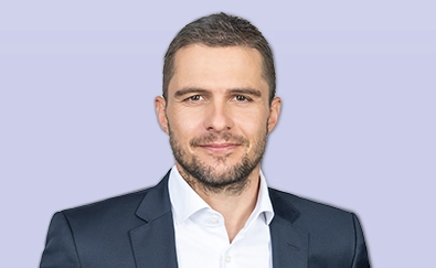 The image shows a person, presumably a man, posing for the photograph. He appears to be a young adult with short hair and some facial stubble. The individual is dressed in formal attire, consisting of a dark suit jacket over a lighter shirt. The background is plain and does not offer any context or additional information about the location or event. There are no visible texts on the image, keeping the focus on the subject of the photograph. The style of the photograph suggests it might have been taken for professional purposes, such as an identification photo or promotional material.