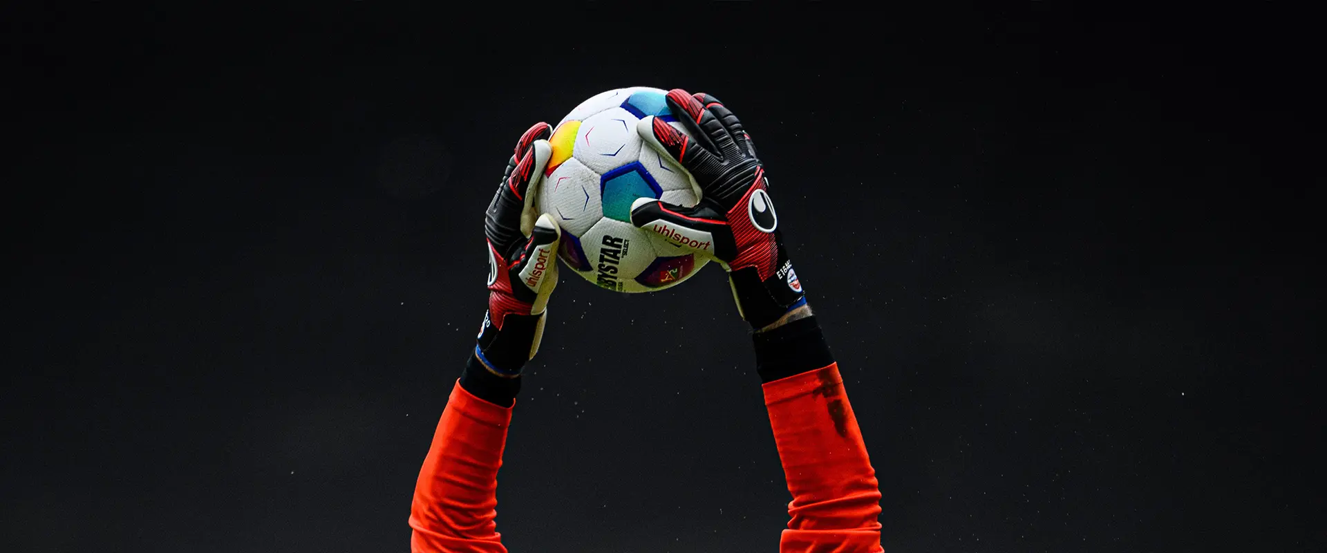 The image shows a hand wearing gloves positioned in such a way that the palm faces downwards with its fingers curled upwards to form a small ball, which is black and white, possibly an abstract or stylized representation rather than depicting a real soccer ball. In the background, there appears to be someone on a pitch, likely playing soccer, wearing what looks like a kit in different shades of red and white. The photo captures the moment from below the action, emphasizing the positioning of the hand and gloves in the foreground, while the rest of the image conveys the context of a sporting event, possibly soccer given the presence of an actual pitch and player's attire.