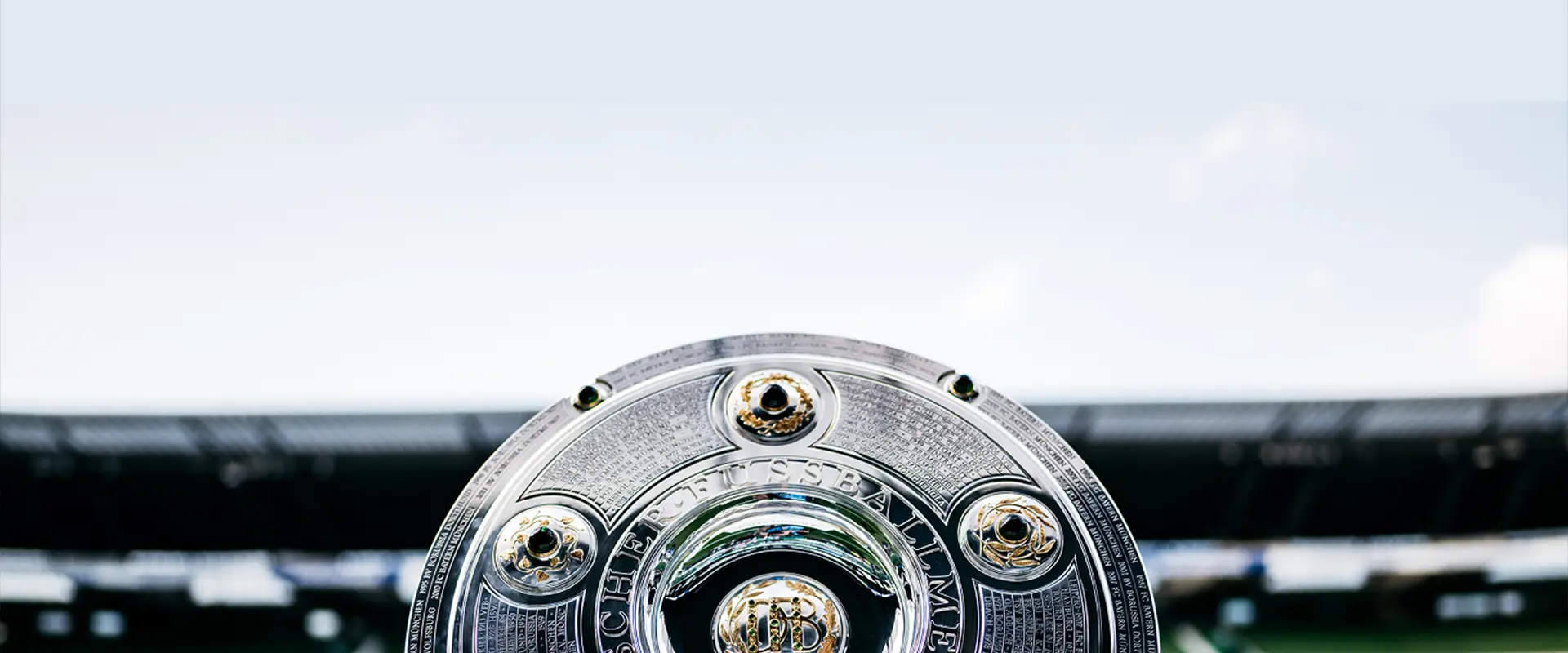 The image appears to be a professional sports trophy, which seems to have multiple stages or cups on its top part. It's set against an out-of-focus background, which makes it difficult to discern specific details about the setting. The trophy itself has a reflective sheen on some parts and is adorned with plaques or badges that could represent logos of sports clubs, teams, or sponsors. There are three visible bands at the bottom, possibly indicating different levels of awards in the competition for which this trophy is presented. The background suggests an event or ceremony where such trophies are typically displayed.