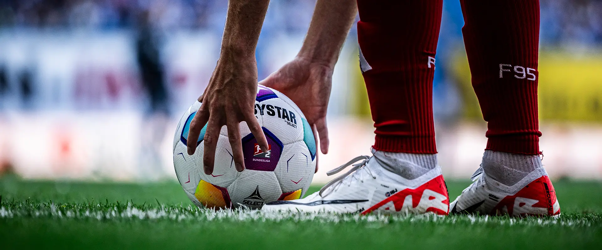 The image shows a soccer player preparing to perform a penalty kick during a football match. The player is focused on the ball, holding it in their left hand while standing with their left knee almost touching the ground, which is common to create a balance point for generating power and accuracy. The player appears ready to make contact with the ball, which is off the ground, indicating that they are about to strike the ball. The footwear, turf, and stadium setting confirm this is an organized professional event. There are no visible texts or brands on the image, and it is a clear, high-resolution photograph capturing the player's action with some context of an ongoing game.