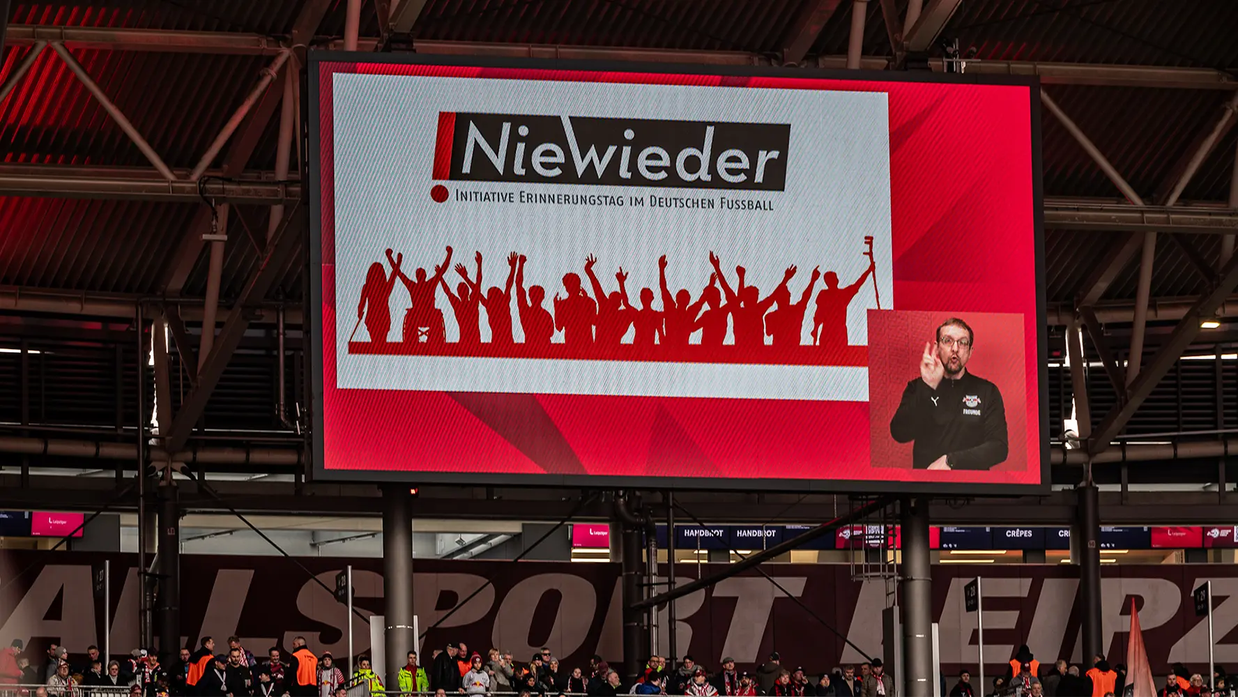 The image shows a large sports event in progress with fans watching from the stands. There's a giant screen displaying the Niewsdeer brand and a person on stage who appears to be speaking to the audience or addressing supporters. Around the television screen, there are multiple people, some of them wearing the same colors as the Niewsdeer team, which suggests they are fans. The atmosphere seems charged with excitement, typical for sports events.