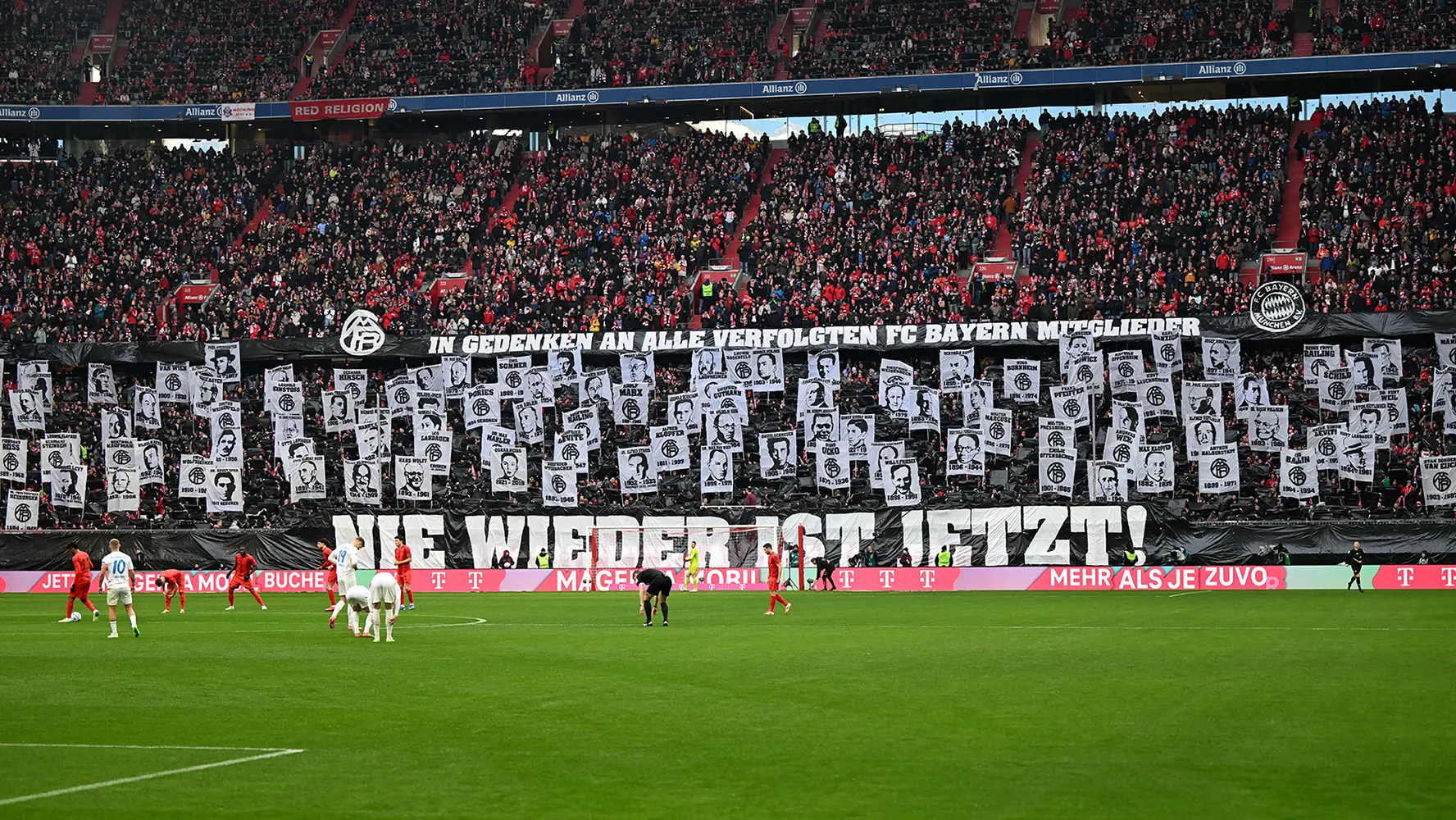 This image appears to show a vibrant and somewhat chaotic scene from an event, likely a sporting match given the presence of football players on the field. In the stands, fans are holding up banners expressing their support or possibly criticism towards different entities or individuals. The text on the banners is in German, suggesting that the venue may be in Germany, and it's clear that these supporters are very passionate about the game or event they are attending. The overall atmosphere is lively and energetic, which is typical at football matches where fans gather to cheer for their team.