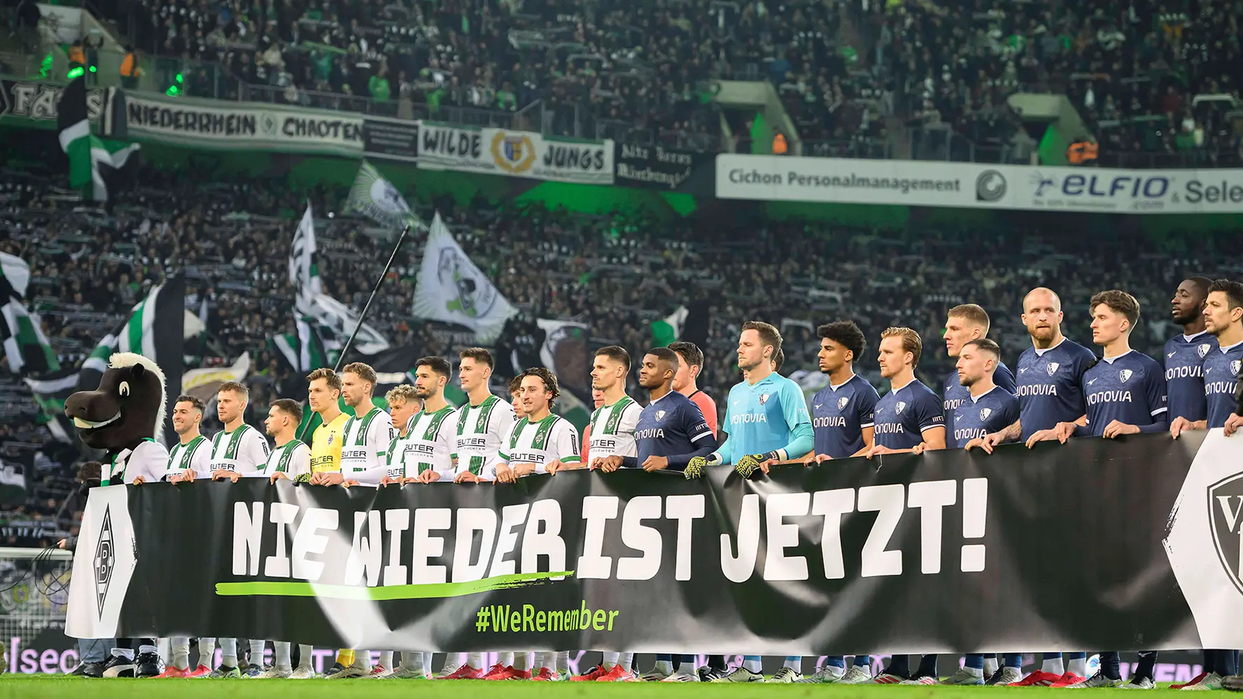 The image shows a soccer team with coaches and players standing on a stage or platform, facing their respective audience. They all appear to be at a formal event, as indicated by the presence of chairs and podiums. There are banners with different logos, suggesting this could be a gathering related to the sports organization or an event that the team is associated with, such as a match dedication, championship celebration, or promotional photo session. The atmosphere seems to be ceremonial in nature.