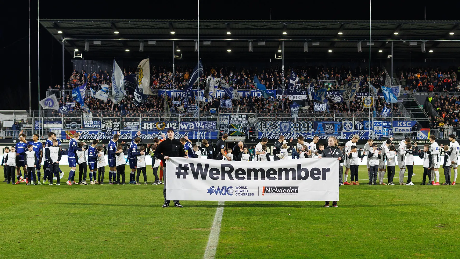 In the image, we see a group of people on a soccer field that is presumably wet due to rain as there's a slight reflection visible on it. The attire suggests the individuals are part of a soccer team or an associated sports event. They are huddled together, possibly in conversation or listening to something. There is an informing sign among them with the words "Welcome home" and "#WEare1," indicating this might be a welcoming or celebratory occasion, such as a return from international play or a homecoming event. The environment suggests this could be in preparation for an upcoming game, given the presence of stadium seats in the background, which are covered but have lights on. The overall atmosphere is suggestive of the end of a match or a special day of celebration associated with the sports team.