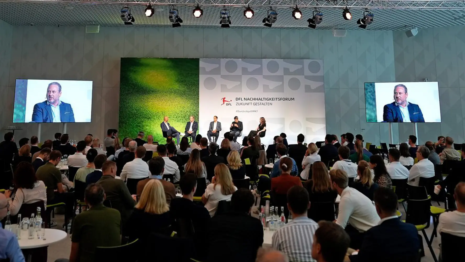 The image displays a conference or event setting, with attendees seated at tables facing a platform with speakers. On the stage, there are three people, two of whom appear to be moderating or speaking at the event. A panel is present on the stage, indicating some form of discussion or evaluation. Behind the panelists, there is a backdrop with promotional displays that include branding and possibly text relating to the event's theme or content. The attendees are dressed in formal or business casual clothing, suggesting a professional audience. Above the room, ceiling lights hang from the ceilings, providing illumination for the space. There is no visible text providing specific information about the event within the image.