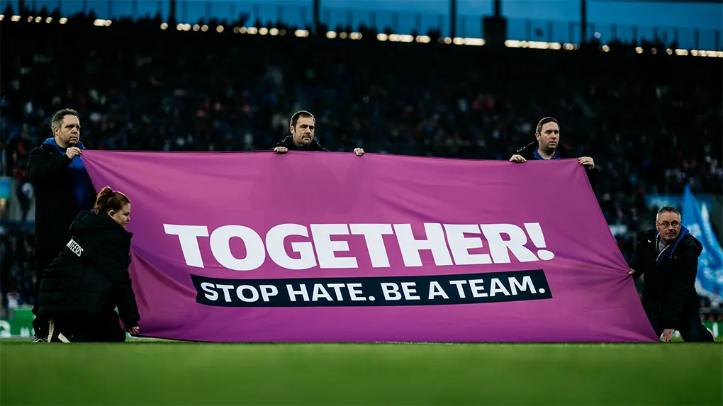 Das Bild zeigt People at a sports event who are protesting against hate speech and racism as part of a campaign to "Stop hate, be a team." It appears that the event is taking place during a soccer game, given the stadium seats, grass field, and players in the background. The flag, reading "TOGETHER STOP HATE BE A TEAM," serves to promote unity and respect.