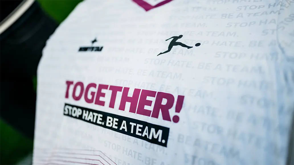 The image shows a team jersey with the number "10" on it, and the name of a team above the shirt. On the chest area, there is sponsor branding. There are slogans such as "TOGETHER WE ARE A TEAM" indicating a team ethos or values statement. The top part of the image shows a bumper sticker with its own message related to inclusivity and stop hate, which appears to be an anti-hate sentiment. The photo seems to have been taken from inside a vehicle, as evidenced by sunlight shining on the jersey's upper left side.