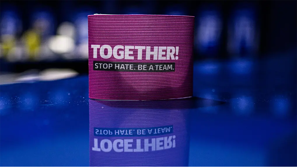 The image shows a pack of playing cards displayed on a reflective surface. In the background, there is text in German that translates to "Together: STOP HATE BECAUSE WE ARE ONE." However, only the second word ("STOP") and the phrase "WE ARE ONE" are visible through the reflection. The image seems to be promoting an anti-hate message and appears to be related to a gaming or entertainment context.