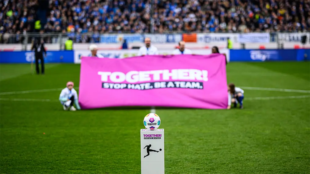 The image depicts a public stadium scene, featuring a banner displaying the message "Together stop hate. Be the change." This suggests that there might be a socially conscious event taking place on this field. The banner is pink, and it stands out on the soccer pitch with the visible blue of the pitch itself around it. In the foreground of the photo, there's an individual squatting down, holding up what appears to be a sign or a flag with additional messaging or imagery, though the text isn't legible in this image. The setting is a soccer stadium during the day under clear skies. There are no visible audiences or spectators, indicating either an early event setup or that the focus of the image was elsewhere, possibly on the banner and the individual with it as representatives of the event's message rather than the event itself.