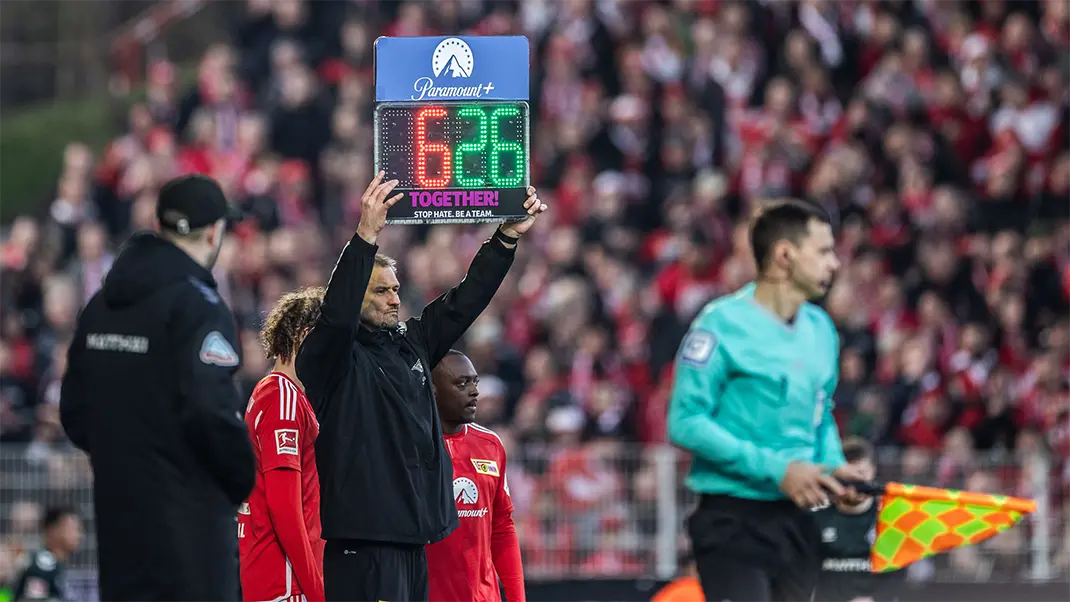 This is a black and white photo showing an event related to soccer, which appears to be a referee holding up a sign with the number "62" on it. The setting looks like a packed stadium, as indicated by blurred crowd members in the background and team personnel or officials in the foreground who seem to be involved in the match or game, given the presence of yellow cards. The overall scene suggests intense competition due to the context of a sports event.