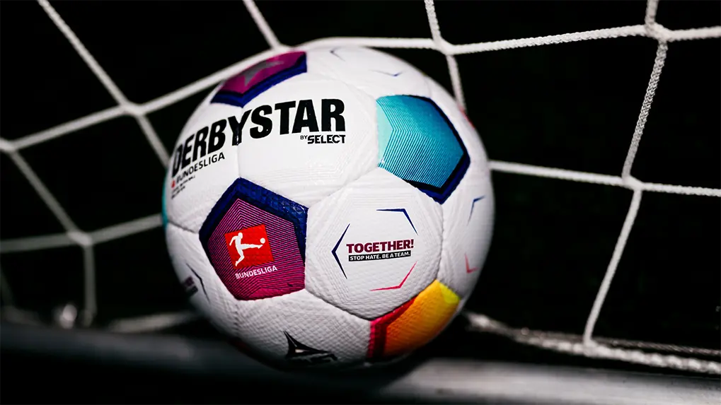 The image you've provided shows a colorful soccer ball placed next to a goalie net or frame, which is characteristic of the sport of football (soccer). The colors on the ball are arranged around a central white pentagon and vary depending on which part of the world the ball is manufactured. This particular design includes elements of stars and stripes, among other patterns. The text "DEBYS" is visible in the bottom left corner, which likely indicates the brand of the manufacturer or sponsor of the ball. The rest of the image features a dark background that contrasts with the vibrant colors of the soccer ball, putting it prominent focus of the photograph.