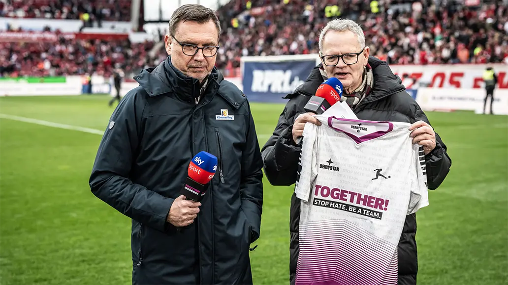 The image shows two individuals at a public event outdoors during the daytime. On the left, there's a person wearing glasses and holding a jersey with a sponsorship logo visible on it. There is a man standing to his right who appears to be interviewing him or engaging in conversation. Both individuals are dressed in informal attire suitable for public events or sports announcements. There is also what seems like a microphone, indicating an event that might be broadcasted or where a speech or announcement is about to take place. The setting suggests this could be a promotional event for a football club or related event.