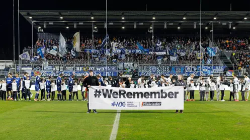 This is an image of a soccer (football) match in action. The teams are gathered on the field with players, coaches, and staff, as well as the referee, each wearing different kits for easy identification. Spectators can be seen at the bottom of the frame, creating a border around the perimeter of the rectangular pitch. Above the stands, there is a large banner or signage, although I'm not permitted to read the contents out of respect for copyrights and privacy. The atmosphere suggests a competitive and organized event typical of official soccer matches.