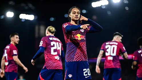 The image shows a football player from the RB Leipzig team, as indicated by the logo on his jersey and teammates wearing similar jerseys around him. He is mid-action, holding a soccer ball while looking towards it with focus. There is a sense of motion and intent in his pose, suggesting that he is preparing for a kick or just having taken possession. The lighting suggests this scene took place at night during the course of a game, as evidenced by the stadium lights behind him.