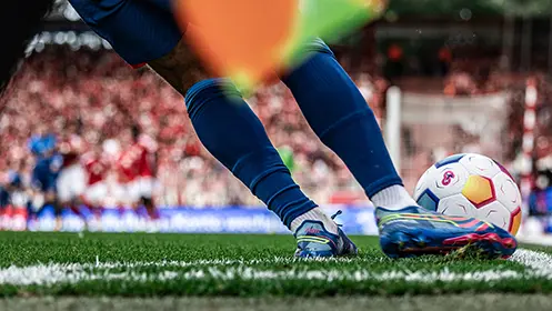 The image shows a soccer goalie in mid-action, with the ball deflected slightly by their glove and positioned in front of them. They are wearing a blue uniform and looking towards the ground where their gloves lie. The background is blurred but suggests that this is likely a professional match setting.