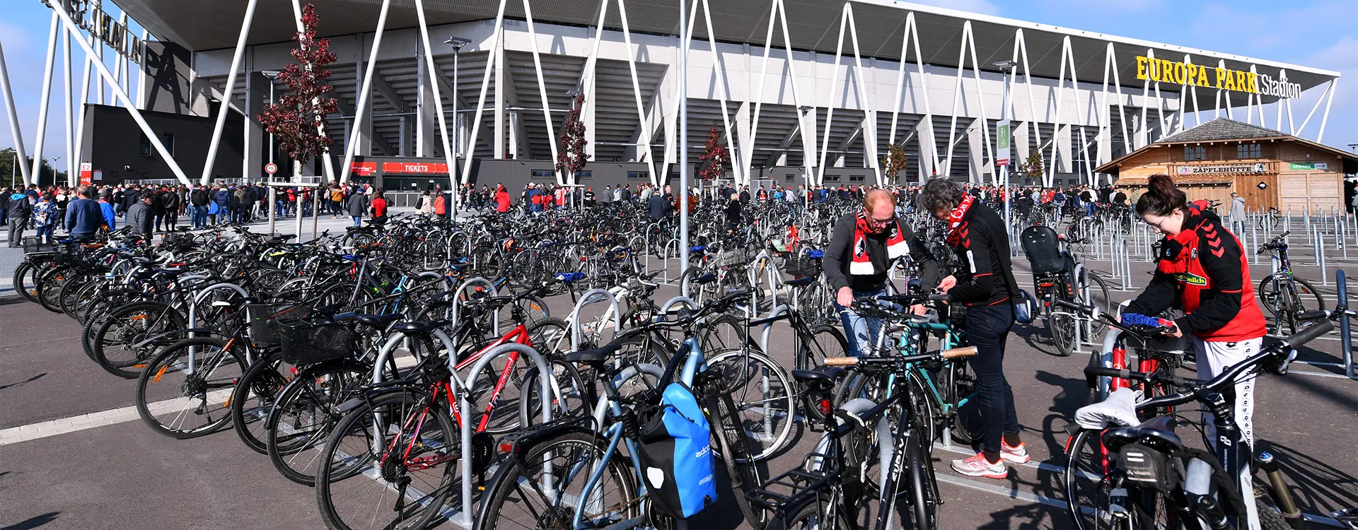 The image shows an outdoor scene centered on a large sports arena with multiple rows of bicycles parked outside. In front of the building, there's a pedestrian sidewalk where people are walking. Overhead, there is some infrastructure which could be part of public transportation or event facilities. It appears to be daytime and the weather looks clear. The surroundings suggest that this might be a special event or gathering for bicyclists, as indicated by the large number of bikes parked in orderly rows.