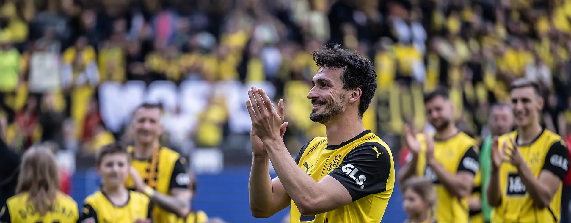 The image shows a person on a soccer field, seemingly interacting with a group of people who appear to be supporters or teammates. This individual is dressed in what looks like a sports jersey and is clapping their hands while standing amidst the others. The background suggests that this is happening at a stadium during a sporting event, as evidenced by the empty seats and the flags in the stands. The focus of the scene seems to be on the person in action rather than any specific detail within the photo.