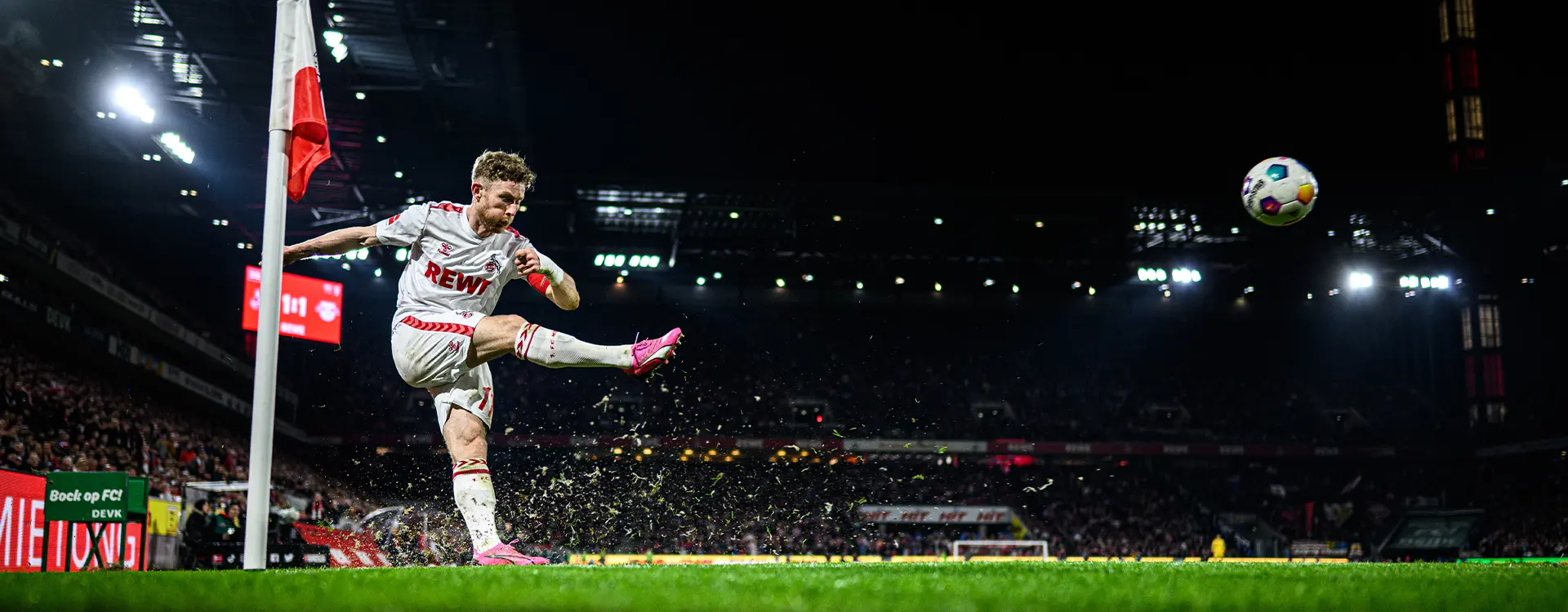 The image shows a soccer player in mid-kick on a football field at night with stadium lights illuminating the scene. A tall goal post is visible to the left of the kicker, and there's a flag in the background suggesting a football match may be underway or about to start. The player appears focused on the ball, which is in motion near their foot.