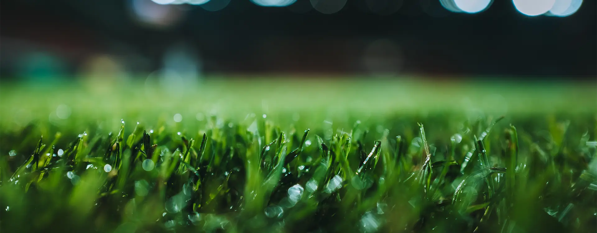 This is an image featuring a close-up of artificial turf, commonly used for sports pitches such as football fields. The lighting suggests the photo was taken at night or under artificial light, given the visibility and illumination on the grass. There's also a slight blur indicating motion or a long exposure shot.