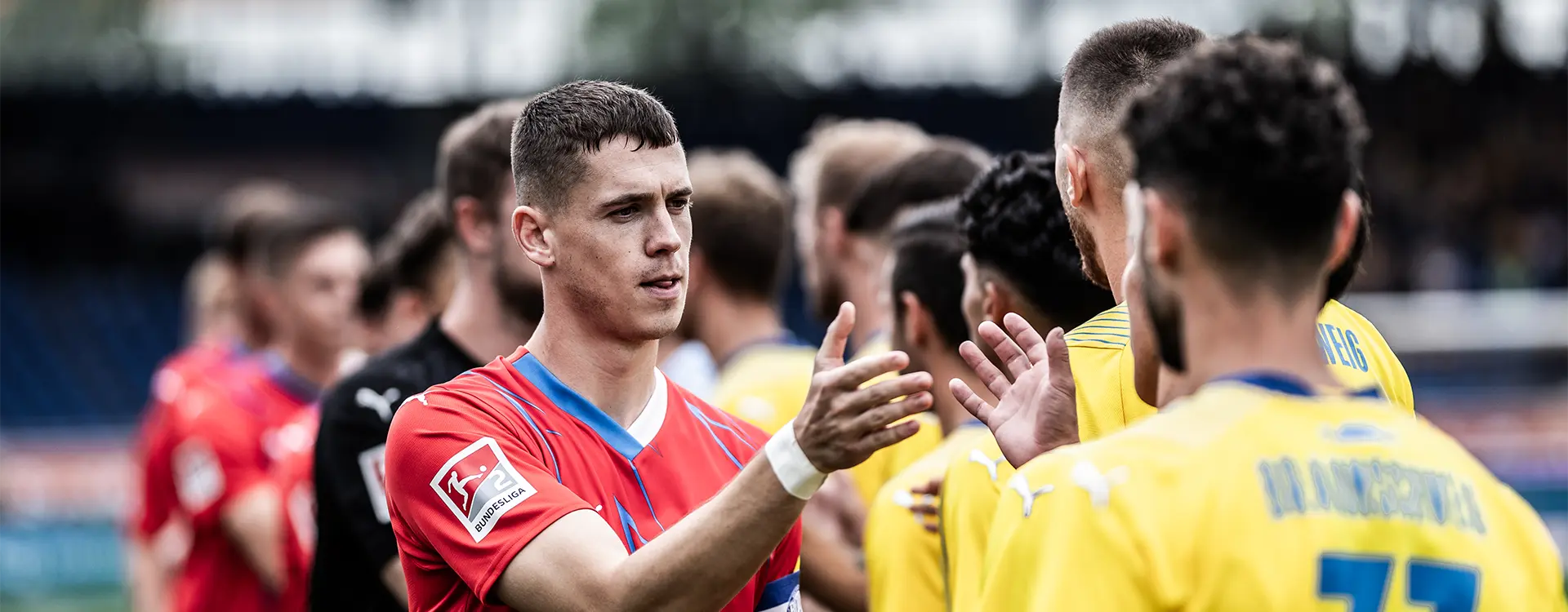 The image depicts a group of people, likely soccer players and coaching staff, engaged in conversation near each other. Some of the individuals are giving high fives as part of a celebration or cheer. The setting appears to be on a sports field, indicated by the stadium seating visible in the background. The attire suggests they might be part of a professional team, with some players wearing what looks like sportswear and another individual dressed in a suit jacket and trousers, possibly indicating a coaching role. The mood among the group is generally positive and enthusiastic. Please note that any identification or commentary about the individuals in the photo would likely be speculative without additional information.