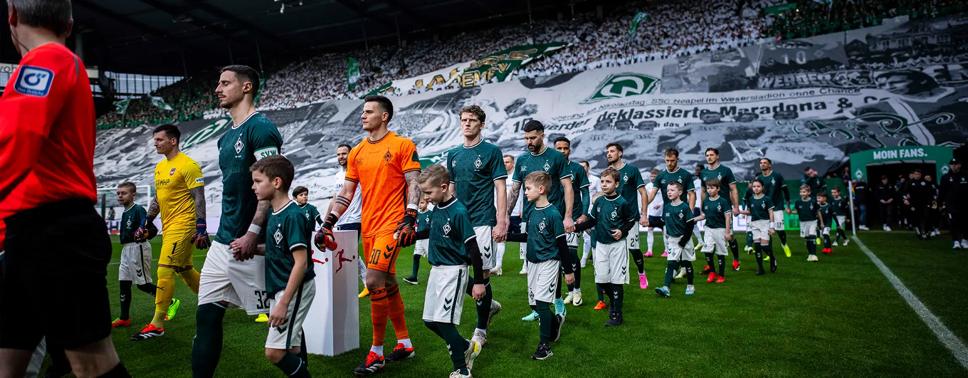 The image shows a group of soccer players assembled on a football pitch. They are dressed in sports gear, which suggests they may be preparing for a match or participating in a training session. The players are positioned behind a long banner that displays what looks like sponsor logos. The setting appears to be during an evening or night event at a stadium, as evidenced by the stadium lights and the darkness visible through the gaps between the benches where players are seated. This implies that the photo might have been taken during a warm-up or before the actual match starts. There is also a scoreboard visible, indicating that this could be a professional stadium.