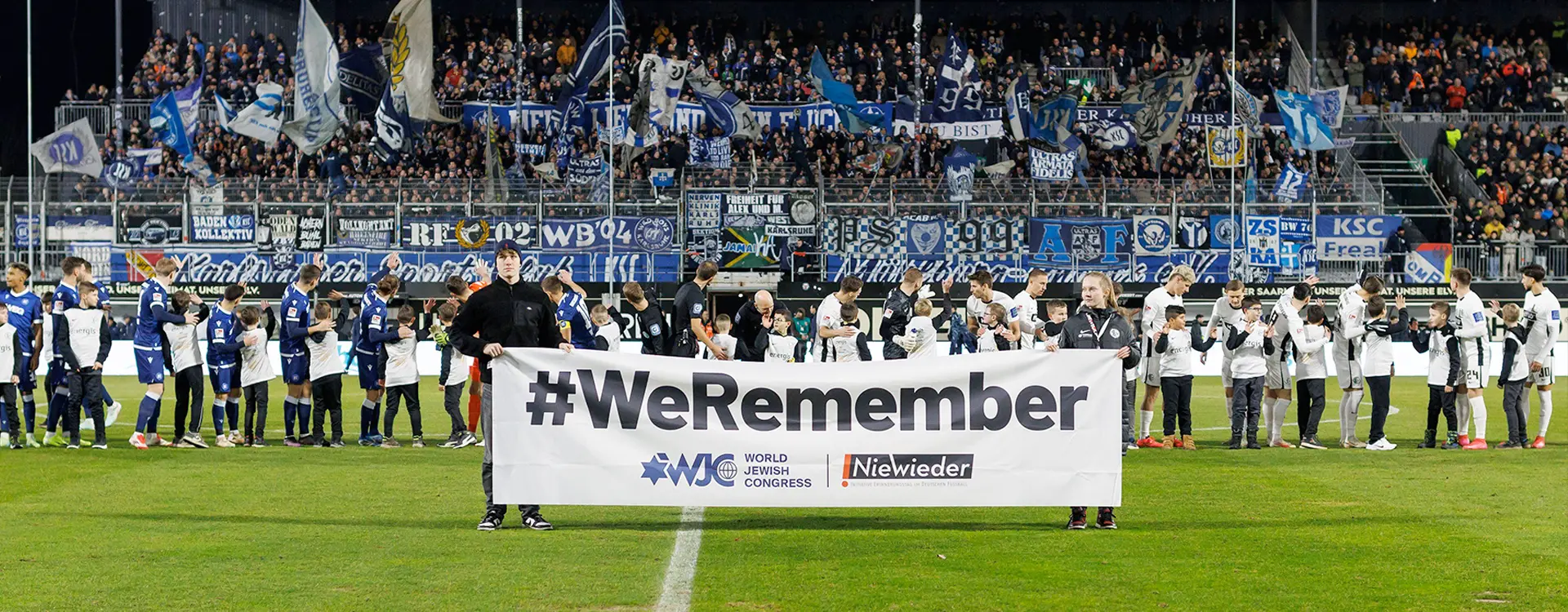 This is an image of a group of people standing on a sports field. They appear to be a mix of professional athletes and possibly team staff. In the background, spectators are visible in a stadium setting with seating visible above them. There's a view over the heads of the people in front where you can see part of the crowd, possibly indicating that event is taking place within a larger sports competition or tournament. The people in the foreground are holding something that says "WE ARE MEMBERSHIP" which could be a slogan or initiative related to the team or organization they represent. There's also a visible flag representing football, suggesting that this event might be related to a footballing match or conference.