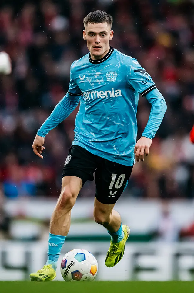 This is an image of a person who appears to be a soccer player. He is wearing a blue jersey with some designs on the sleeves, which suggests he might be part of a sports team. The player is in motion, kicking the ball, which indicates that he is actively engaged in playing soccer. The background shows what seems to be a stadium with rows of seating, suggesting this photo was taken during an actual match or training session. The image also features text at the bottom, but it's not readable in the provided resolution.