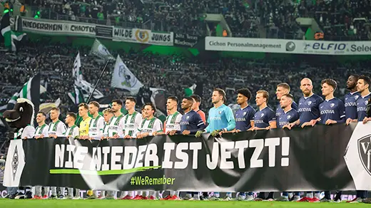 The image shows a soccer team presented on a banner with two other smaller banners underneath it. The central banner is the most prominent and features the photo of the players from a sports club, along with the name of their local municipality "Mönchengladbach." There are also logos for sponsors and what appears to be an advertising slogan or message.
Two additional banners can be seen at the bottom of the main one; they seem to be promotional materials for events or games in this city. It has become a common practice after soccer matches where local teams celebrate their successes on the field, as evidenced by the banner "WEI DEUTSCHLAND IS WORLD CHAMPION" at the bottom of the larger one, indicating the pride and support for the national team's victory in the 2022 FIFA World Cup.
The setting looks like a stadium with a crowd visible in the background, suggesting this is a post-match scenario where the local club's players are posing on the field with fans behind them. The atmosphere appears festive and celebratory due to the accomplishment of their national team.