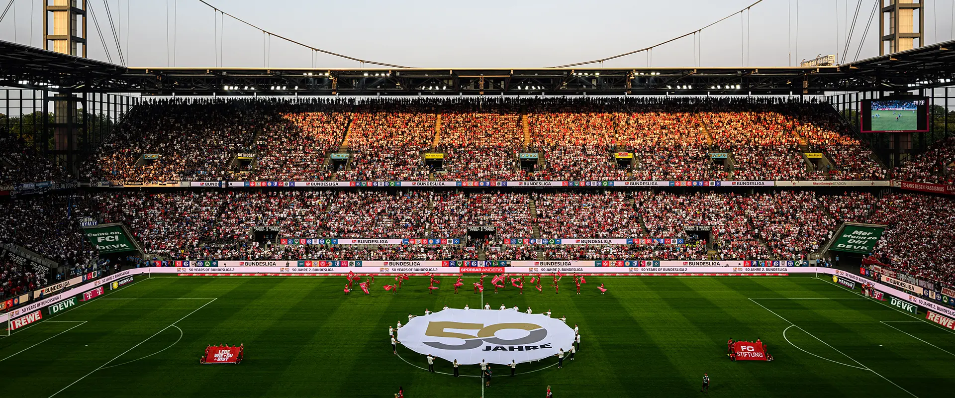 The image shows an aerial view of an outdoor football stadium during daylight. Spectators can be seen in the stands, with some sections of seating filled by people who appear to be watching an ongoing game. The pitch is visible, marked for the sport and with the goalposts aligned vertically. There are no distinct objects or elements that identify the location other than the typical features found in such a setting. It's impossible to determine the event taking place from this perspective.