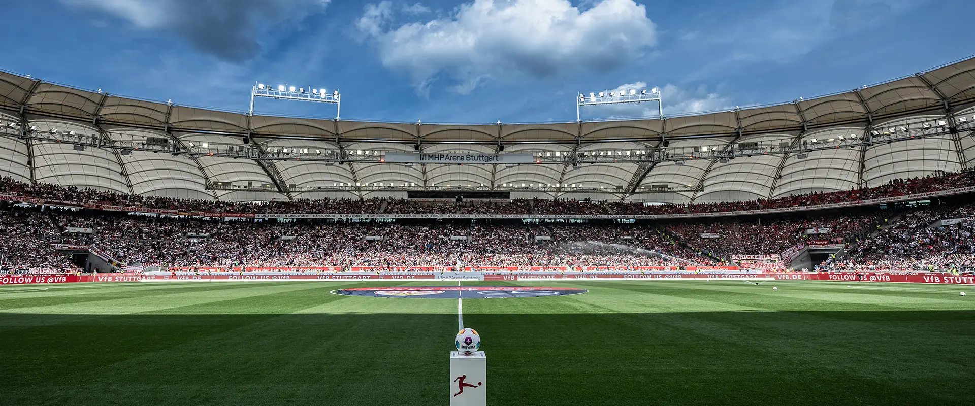 This is an image of an empty stadium during the day. The seats are arranged with white panels on the railing, indicating it might be a football or soccer stadium. In the foreground, there's a grassy field with clear skies above. The photo seems to have been taken during the day when there isn't any event taking place in the stadium.