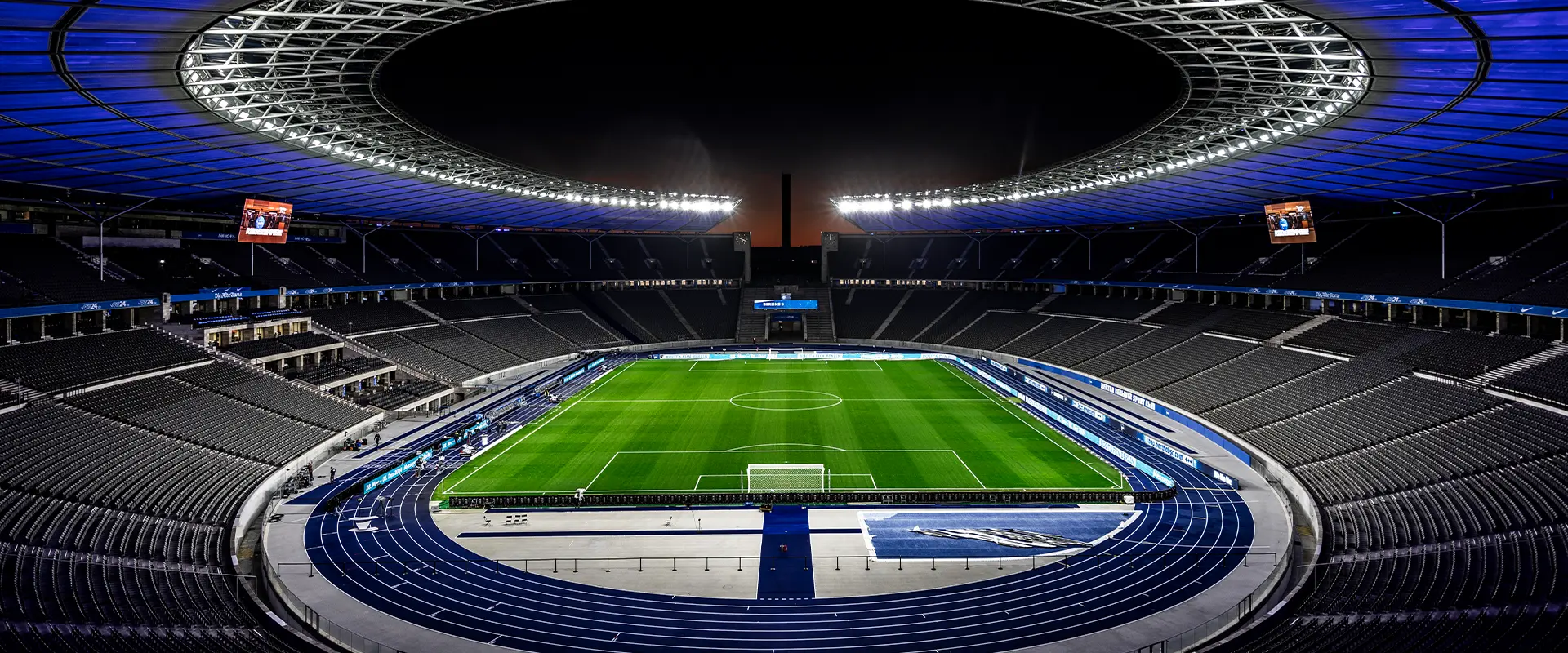 The image shows a nighttime view of a stadium interior with the stadium seats visible from the perspective of the stands. A large, curved arch or roof structure is over the seating area, suggesting it's an indoor stadium with a retractable roof. There are white lighting fixtures mounted on this arch. The photo captures the wide expanse of empty seats in the absence of spectators, which gives a sense of scale and emptiness. The artificial turf playing surface is also visible and marked out for sports like soccer or rugby. The surrounding areas have some temporary seating sections with benches for spectators who are not present in this moment. The lighting creates an impressive atmosphere, and it appears to be a well-maintained stadium.