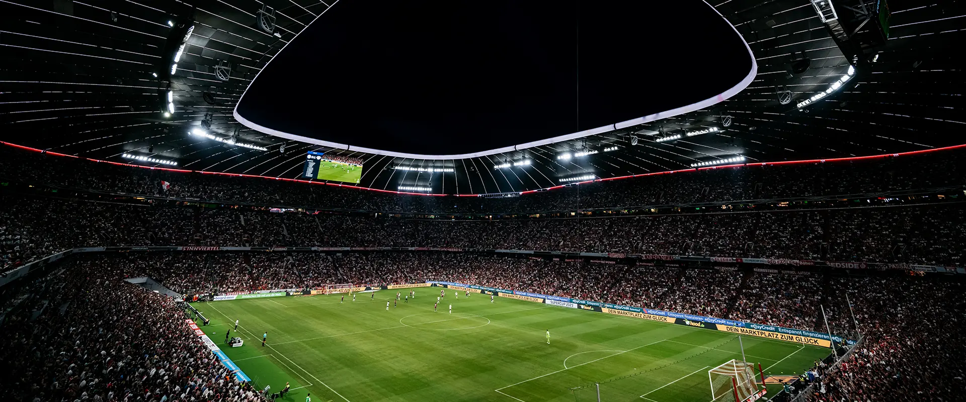 This is a photograph showing an interior view from one of the stands inside a large sports stadium, which appears to be during a football (soccer) match. The focus is on the central area of the pitch with lines drawn indicating areas of the field where out-of-bounds situations occur. The lighting indicates it's night time, and you can see an audience in the stands above that portion of the photo where the lighting is visible, watching the game. There are no spectators in view due to the angle of the image, taken from below the seating areas.
The photograph has been altered or edited to include a watermark that reads "WATCH," which may indicate the source of the image or could serve as a branding for an entity involved with watching sports online. The watermark is semi-transparent and does not detract from the main content of the stadium's interior and the layout markings on the pitch.