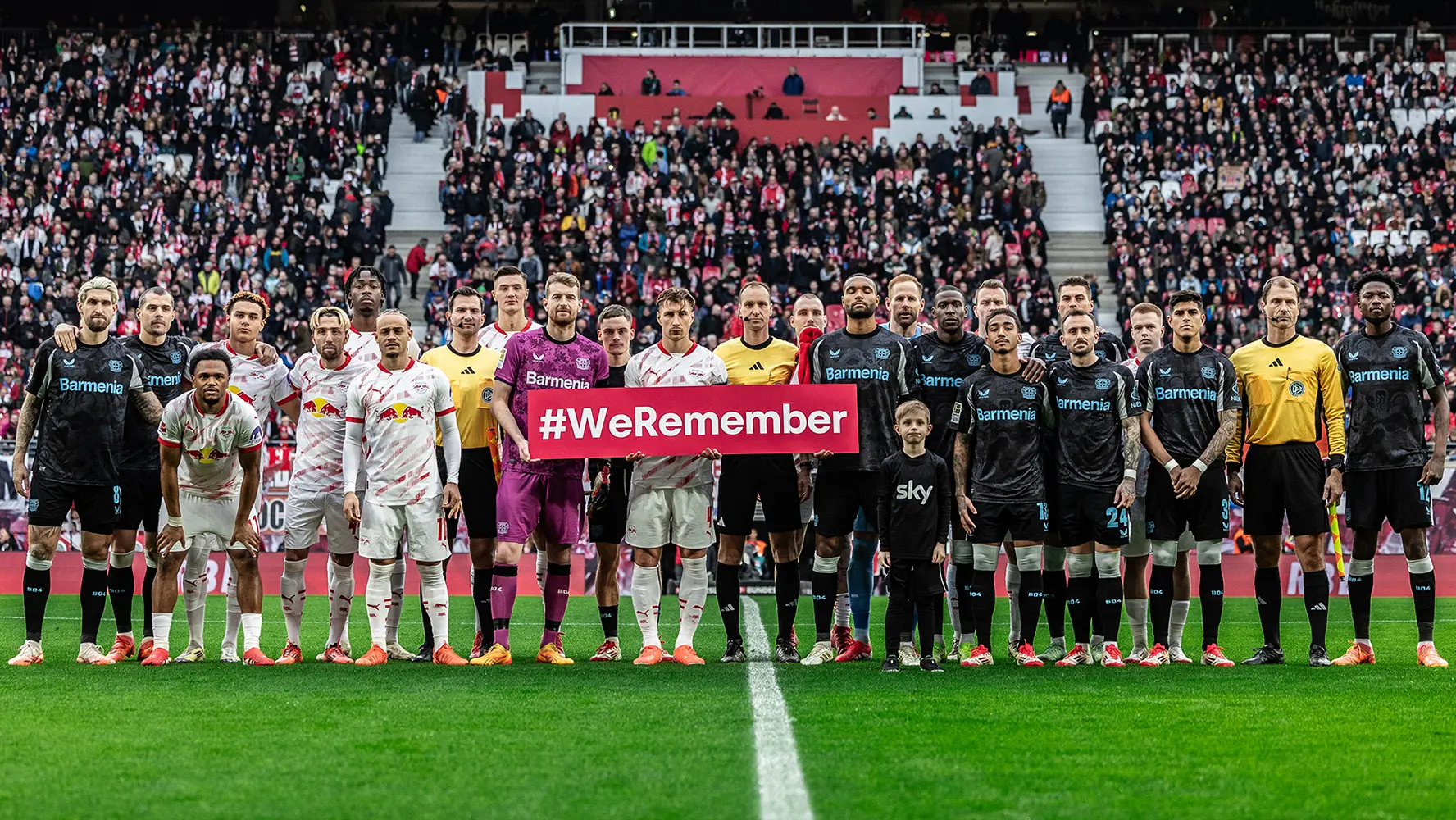 The image shows a group of soccer players on a field. They are standing in front of what appears to be the goal, with some soccer posts and a "WHERE EAGLES LAND" sign nearby, which is likely an advertising slogan for the venue or event. In the background, there are empty stands showing only a few spectators present, suggesting that this photo might have been taken before a match starts or after it ends. The players are arranged in two rows; the front row is standing closely together with their hands on their hips, while the back row is slightly apart and looking towards the crowd. They all seem to be posing for the photo, though one player has his focus directed off the field to his side. The atmosphere appears to be formal and organized, in contrast to the action typically associated with a soccer match.