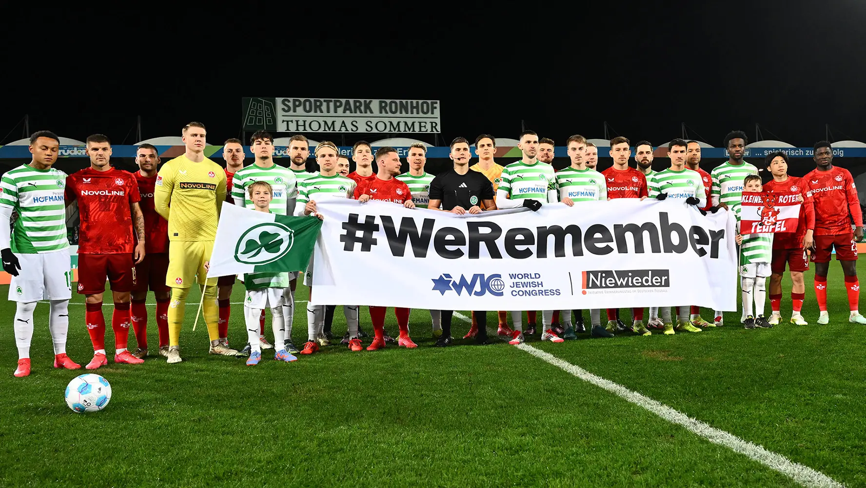 The image shows a football (soccer) team posing with what appears to be a banner. The text on the banner reads, "WE REMEMBER," and there's an Irish flag above it. The team members are wearing football kits with various sponsors visible. The environment suggests this is either at their home stadium before a match or at an informal event related to the sport.