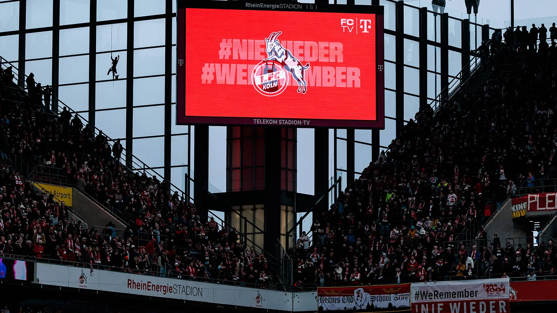 The image you've shared is a photo taken from the stands at a sports event, specifically an indoor stadium. There is a large screen in front of the camera where there appears to be some text and possibly a video or live feed that's not clear from this viewpoint. Below the screen, the area is crowded with spectators. The atmosphere suggests that a significant event is taking place, with people gathered to support the teams or watch the game. The setting looks professional, likely indicative of a soccer or other football match given the screen and the context provided by the content on it." #Wieder" is visible on the display, which might have been intended for the scoreboard but in this image, it seems to be part of a message or banner being displayed. The setting looks like it's either at the start or during the match, given that there is no play happening and the spectators are not engaged with the ongoing action.