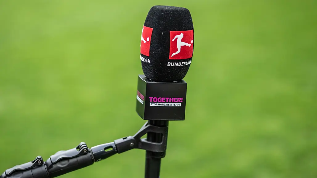 The image shows a microphone with red accents, placed on top of what appears to be an electronic device. This setup is commonly used for broadcasting events such as sports commentating or live news reporting. The background is out of focus, emphasizing the microphone and audio equipment. It suggests that this image may have been taken during a broadcast from a studio or the location where the person is discussing or calling an event such as soccer.