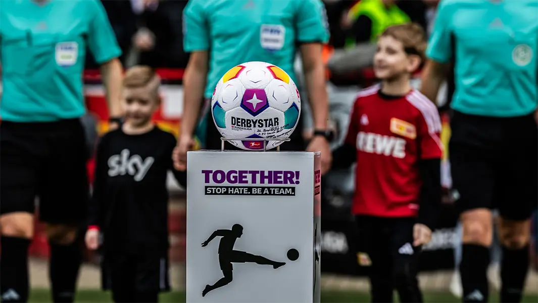 The image shows a group of people on what appears to be a soccer field, with soccer balls and a sign that says "Together we are Stronger." In the foreground, there's a large ball mounted on top of poster board. There's also text "3RSY" on the posters behind the participants. The setting suggests this might be an event or gathering related to sports, perhaps for a charitable cause or team sponsorship.