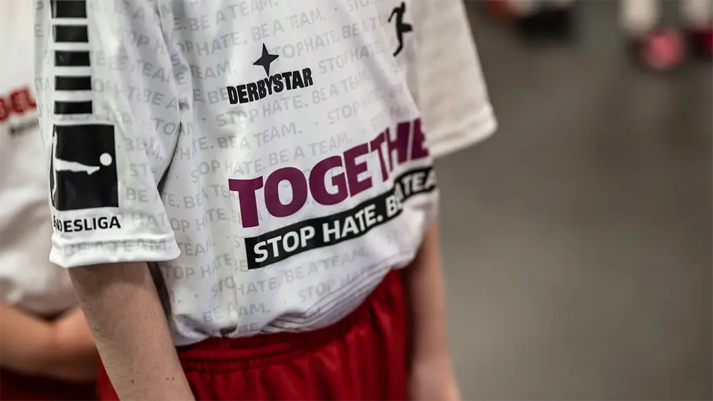 This is an image of a young person wearing a white and purple sports jersey with the word "STOPTHEHATE_FUTURES" across it. The individual has short hair, possibly parted in the middle, and their face appears contemplative or focused on something out of frame. The background suggests that this photo was taken at a sports facility or sports event due to the presence of football pitches. There is text visible on the jersey and presumably on other elements of the facility, but it is partially obscured, making it difficult to read in full detail.