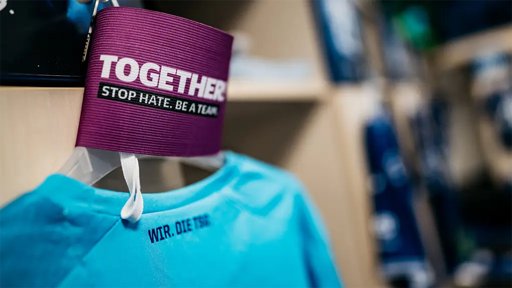 The image shows a clothing item hanging from the top, attached to a hanger. The clothing is predominantly blue and the text "Together - Stop hate, be love" is visible on the garment. In the background, there appears to be clothing display, as suggested by other items of clothing and what looks like shelves or a rack for more merchandise. The setting suggests a store or a market stall where these clothes are being displayed or sold.